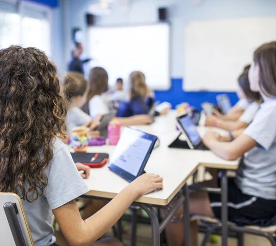 Schülerinnen sitzen im Klassenraum mit Tablets am Tisch und schauen zur Tafel und zur Lehrerin.