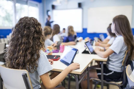 Schülerinnen sitzen im Klassenraum mit Tablets am Tisch und schauen zur Tafel und zur Lehrerin.