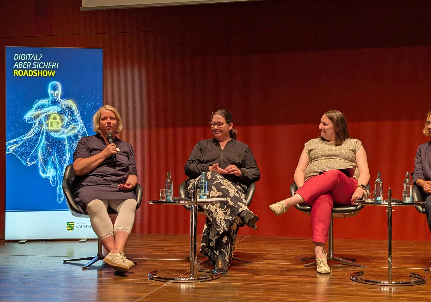 Dr. Juliane Hundert sitzt beim Podiumsgespräch mit anderen Gesprächsteilnehmerinnen auf einer Bühne. Im Hintergrund steht ein Rollup mit der Aufschrift »Digital, aber sicher! Roadshow«.