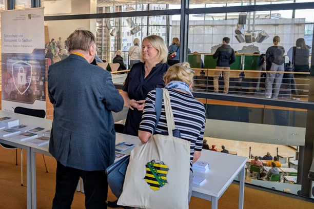 Dr. Juliane Hundert spricht an ihrem Stand mit einem Besucher und einer Besucherin beim Tag der offenen Tür im Sächsischen Landtag