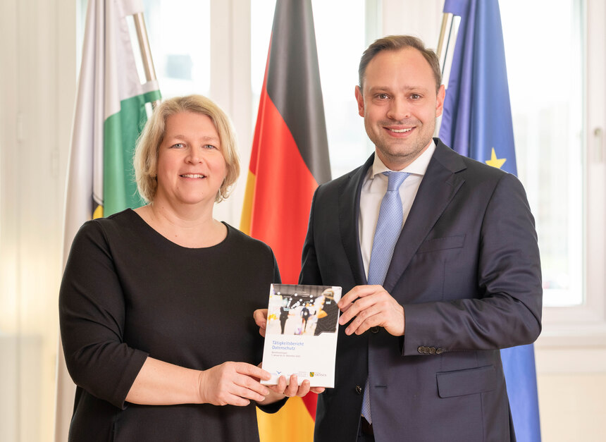 Dr. Juliane Hundert und Alexander Dierks stehen vor den sächsischen, deutschen sowie europäischen Fahne und halten den Tätigkeitsbericht Datenschutz 2025
